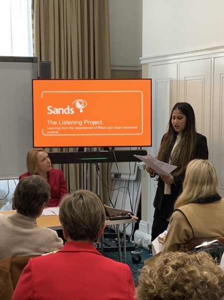 A person speaking to a group of people in parliament in front of a screen that reads 'Sands Listening Project'